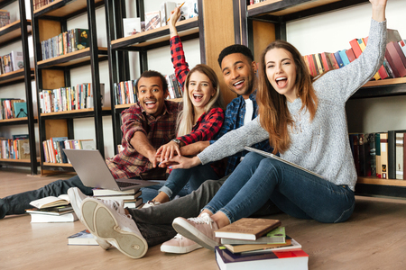 Image of young smiling students sitting in library on floor using laptop computer. Looking at camera.の写真素材