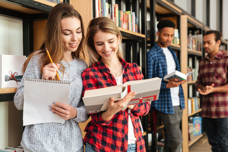 Photo of young smiling women students standing in library reading books. Looking aside.の写真素材