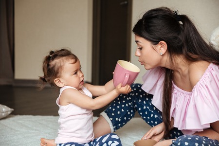 Picture of pretty young mother drinking tea with her little daughter sitting on bed indoors. Looking aside.の写真素材