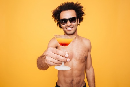 Picture of handsome young african man standing isolated over yellow background. Looking at camera drinking cocktail. Focus on cocktail.の写真素材
