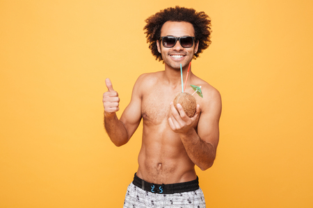 Picture of smiling young african man dressed in shorts standing isolated over yellow background. Looking at camera drinking cocktail and showing thumbs up.の写真素材