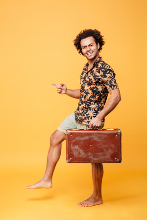 Image of attractive young african man walking over yellow background and holding suitcase. Looking at camera.の写真素材