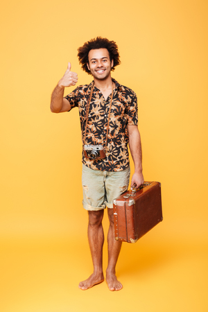 Full length portrait of a happy joyful african man holding suitcase and showing thumbs up isolated over yellow backgroundの写真素材