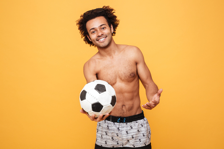 Portrait of a young smiling african guy in summer clothes holding foot ball over yellow backgroundの写真素材