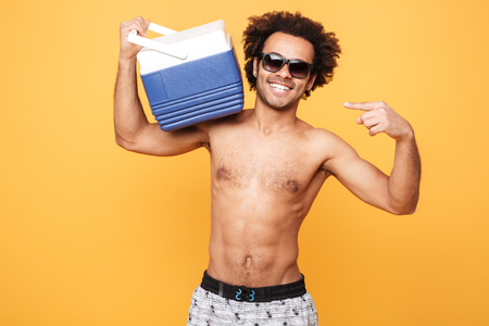 Portrait of a young afro american man in summer shorts holding and pointing at icebox over yellow backgroundの写真素材