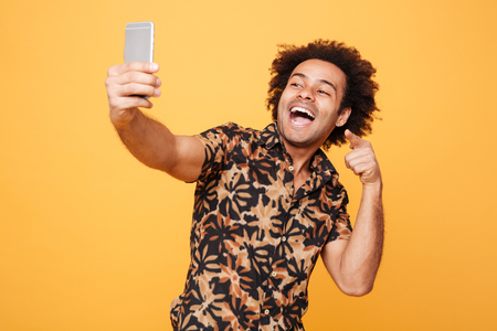 Picture of happy young african man standing over yellow background and make selfie while pointing. Looking aside.の写真素材
