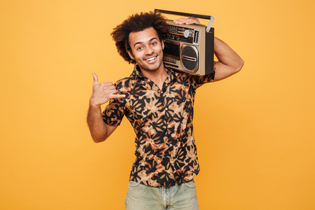 Portrait of a smiling african man in summer clothes holding boombox on his shoulder isolated over yellow backgroundの写真素材