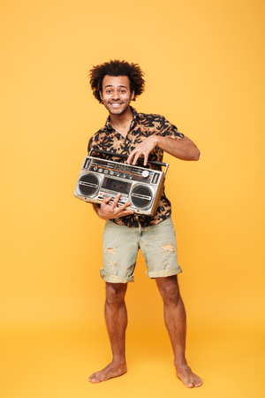 Image of young smiling african man standing with tape recorder isolated over yellow background. Looking at camera.の写真素材