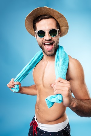 Image of happy man wearing summer clothes and sunglasses holding towel on shoulders isolated over blueの写真素材