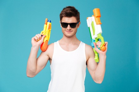 Positive young handsome brunette man in sunglasses looking camera and holding two water guns...