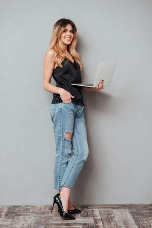 Full length portrait of a smiling pretty woman standing and holding laptop isolated over grey backgroundの写真素材