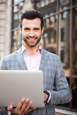 Portrait of a smiling handsome man in jacket using laptop computer and looking at camera while standing in a city areaの写真素材