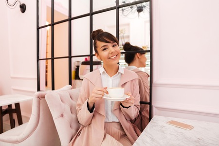 Portrait of a smiling young girl in pink coat drinking coffee while sitting inside a cafeの写真素材