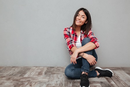 Picture of smiling young asian woman isolated over grey wall sitting. Looking at camera.の写真素材