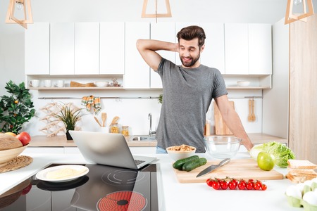 Confused handsome man cooking vegetable salad in kitchen with laptopの写真素材