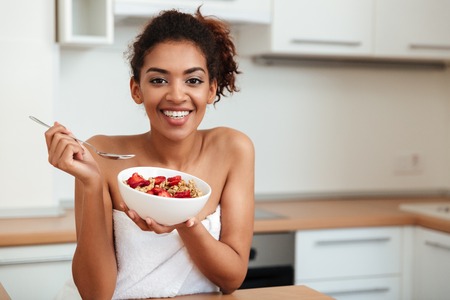 Image of cheerful young african woman dressed in towel sitting at home and looking at camera while eating strawberry with muesli.の写真素材