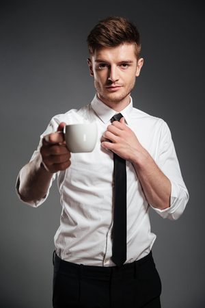 Handsome confident young businessman in formalwear holding cup of coffee and looking at camera isolated over grey backgroundの写真素材