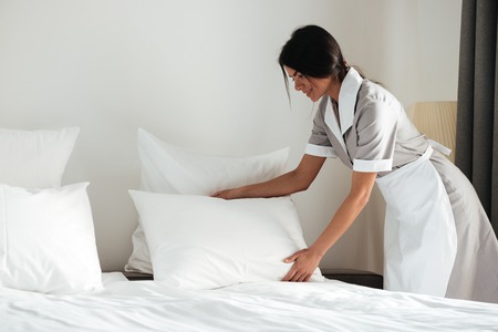 Young hotel maid setting up white pillow on bed sheet in hotel roomの写真素材