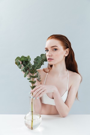 Fashion portrait of a young pretty redhead girl wearing swimsuit holding eucalyptus while sitting at the table and looking at camera isolatedの写真素材