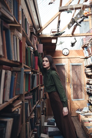 Side view of brunette woman taking book from shelf and looking camera in libraryの写真素材