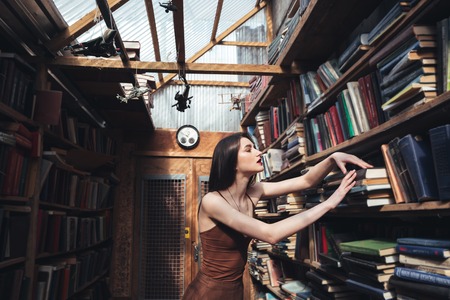 Side view of young brunette woman standing in library and leaning on shelf with booksの写真素材