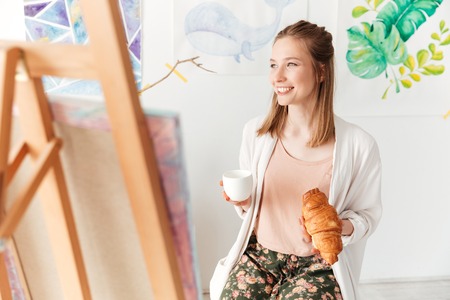 Picture of happy young caucasian lady painter at workspace drinking coffee eating croissant. Looking aside.の写真素材