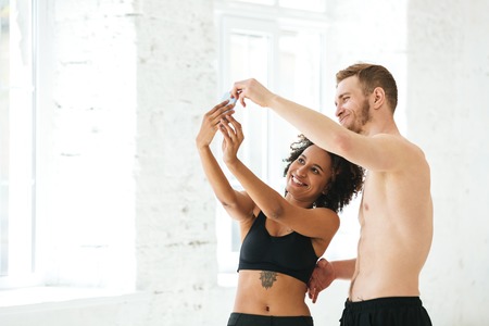 Multicultural smiling sport couple making selfie on mobile phone while standing indoorsの写真素材