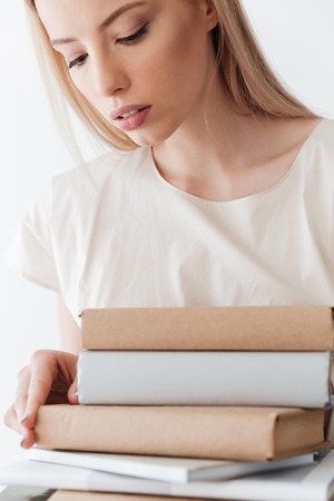 Picture of serious blonde woman standing isolated white background holding books. Looking aside.