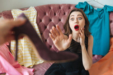 Portrait of a screaming pretty woman buyer in dress sitting on a couch and trying to reach high heel shoes indoorsの写真素材