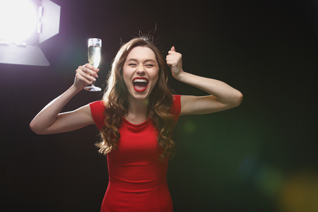 Happy excited young woman with glass of champagne standing and shouting over black backgroundの写真素材