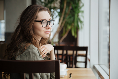 Photo of young happy lady with long hair wearing glasses sitting in cafe. Looking aside.の写真素材