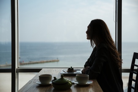 Horizontal shot of young woman enjoying the sea in window while eating in cafeの写真素材