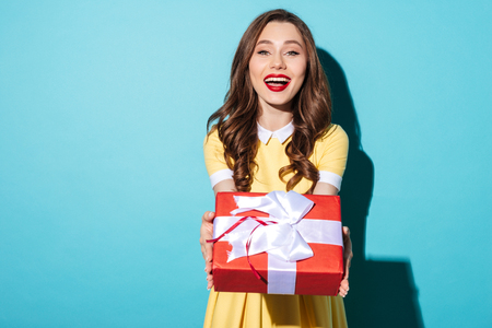 Portrait of a cheerful pretty girl in dress giving present box to camera isolated over blue backgroundの写真素材