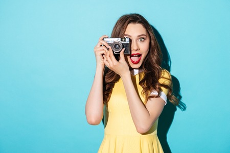 Portrait of a happy cheerful girl in dress using retro camera isolated over blue backgroundの写真素材