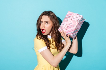 Portrait of an astonished pretty girl in dress holding gift box isolated over blue backgroundの写真素材