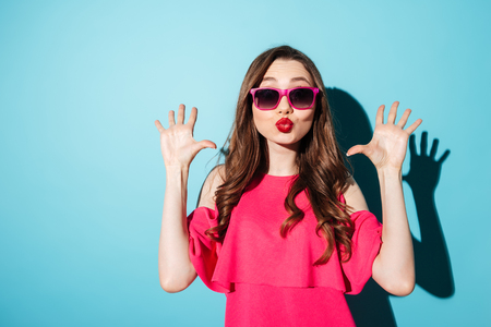 Photo of a beautiful surprised young brunette woman in pink dress wearing sunglasses posing and looking at camera over blue background.の写真素材