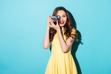 Portrait of a smiling pretty girl in dress taking photo on a retro camera isolated over blue backgroundの写真素材