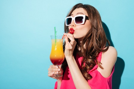 Close up portrait of a cute brunette girl in sunglasses and dress drinking cocktail drink with a...