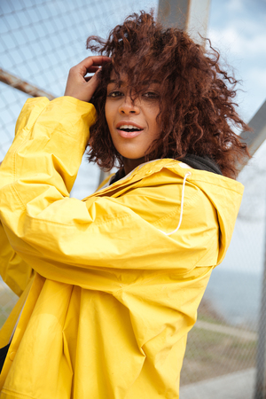 Image of happy african curly young lady wearing yellow coat walking outdoors. Looking at camera.の写真素材