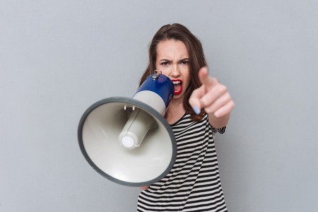 Photo of screaming young pretty lady standing over grey wall holding loudspeaker. Looking at camera and pointing.の写真素材