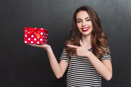 Picture of happy young lady standing over grey wall and holding gift in hands. Looking at camera and pointing.の写真素材