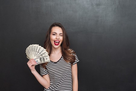 Image of happy young lady standing over grey wall and holding money in hands. Looking at camera.