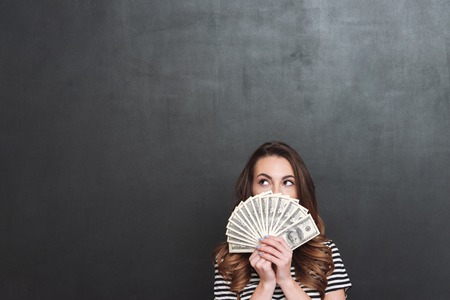 Picture of young lady standing over grey wall and holding money in hands. Looking aside.の写真素材