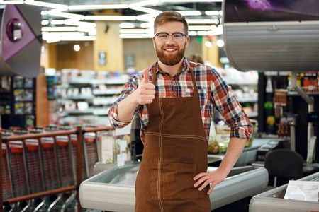 Picture of happy cashier man on workspace in supermarket shop. Looking at camera showing thumbs up.の写真素材