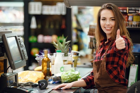 Picture of cheerful cashier woman on workspace in supermarket shop. Looking at camera showing thumbs up.の写真素材