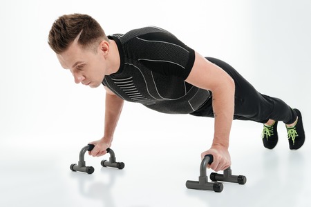 Portrait of a confident healthy sportsman doing push ups with gym equipment isolated over white...