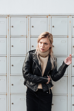 Fashion portrait of angry young blonde lady standing near safes. Looking aside holding cigarette.の写真素材