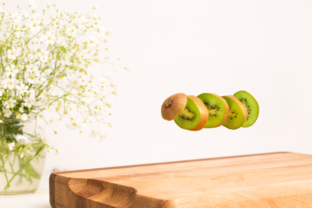 Sliced whole kiwi flying above a wooden chopping board with vase of flowersの写真素材