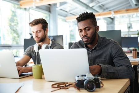Image of serious young colleagues sitting in office coworking using laptop computers. Looking aside.