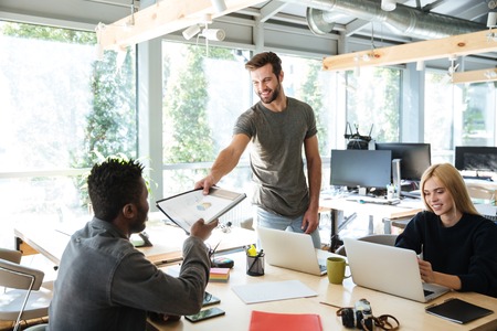 Image of smiling young colleagues sitting in office coworking using laptop computers and work with...